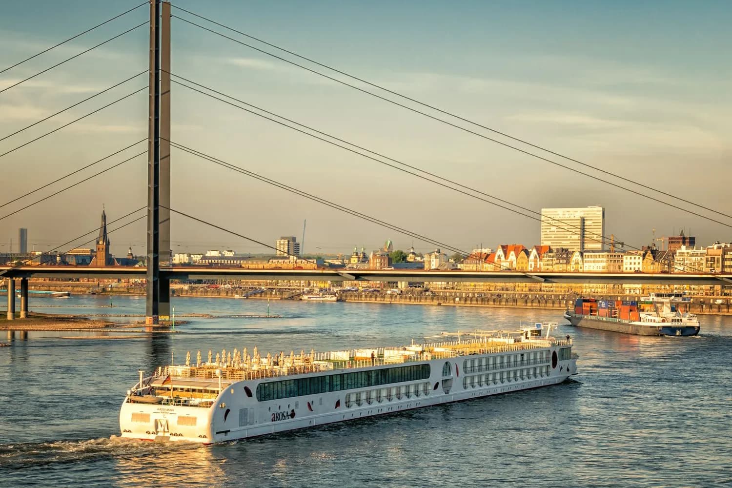 Flusskreuzfahrtschiff fährt auf dem Rhein vor der Düsseldorfer Skyline mit Brücke im Abendlicht