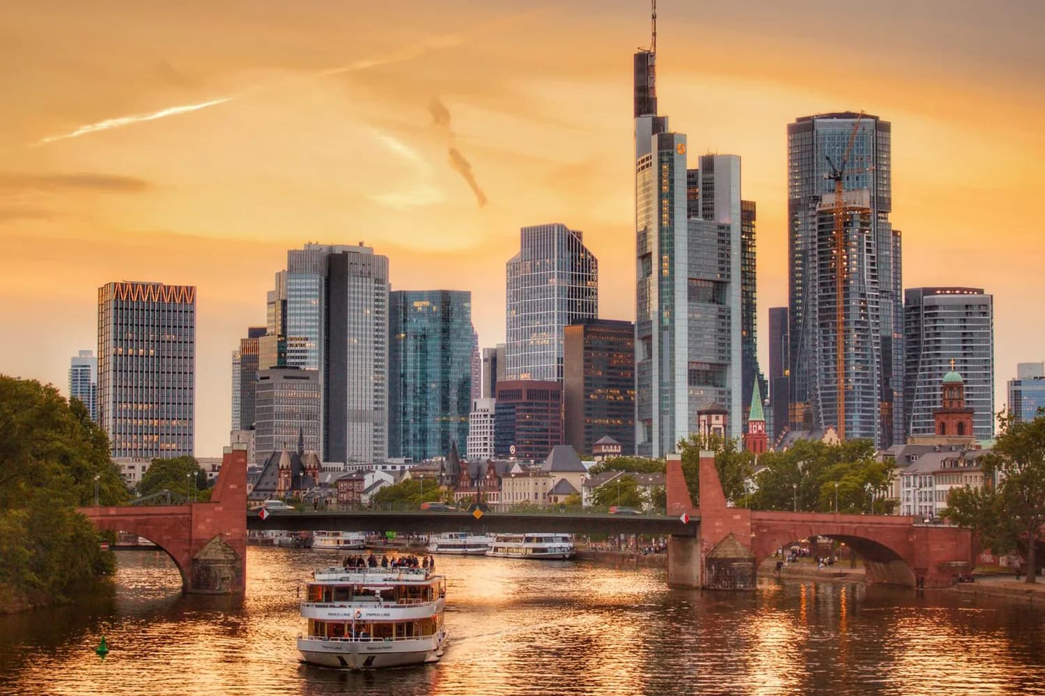 Flusskreuzfahrtschiff auf dem Main vor der Frankfurter Skyline bei Sonnenuntergang mit Brücke im Vordergrund