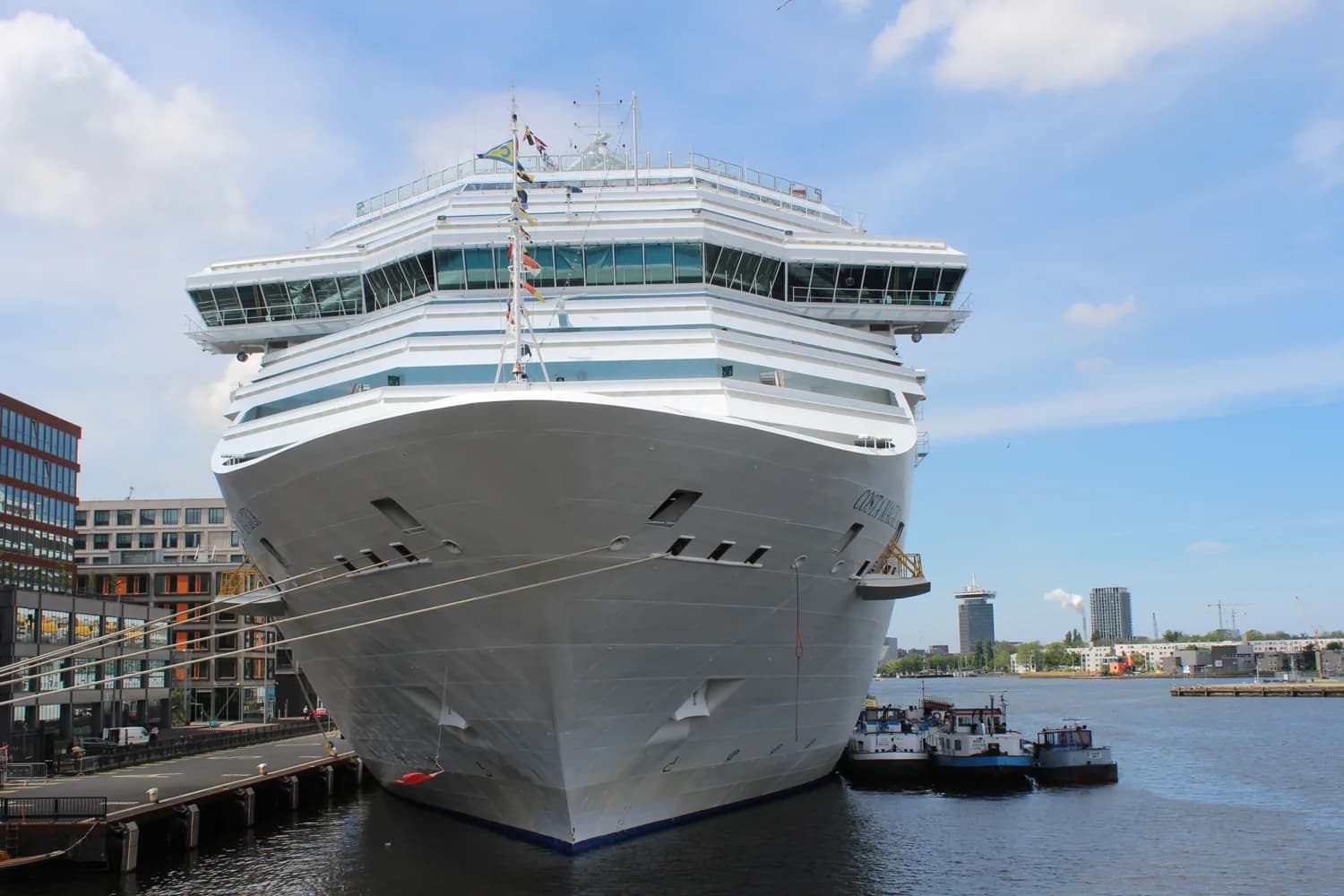 Weißes Kreuzfahrtschiff liegt am Kai im Hafen von Amsterdam, daneben moderne Gebäude und kleinere Boote
