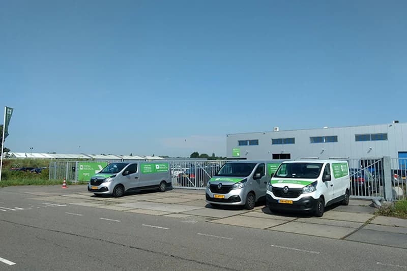 Drei weiße Transporter mit grünen Aufklebern vor geschlossenem Metalltor eines umzäunten Betriebsgebäudes am Hafen Amsterdam.