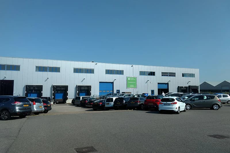 Parkende Autos vor großer Lagerhalle mit blauen Ladeöffnungen und grünem Schild bei klarem Himmel.