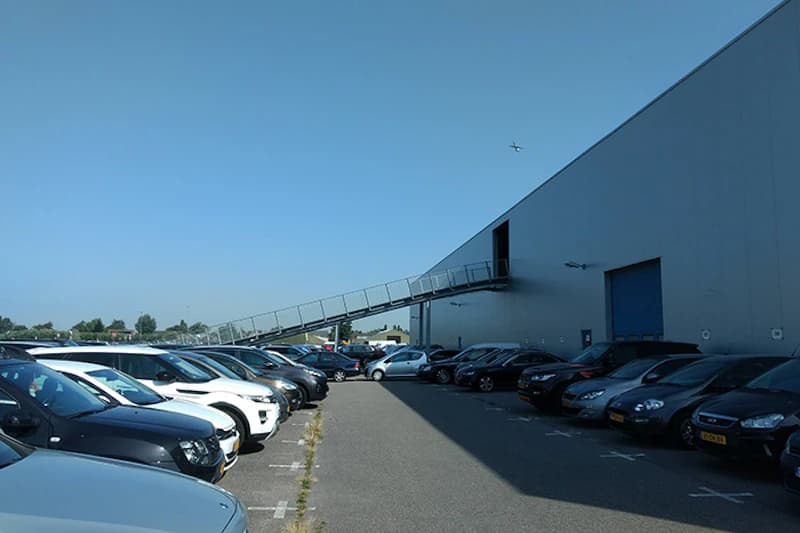Außenparkplatz neben großer Halle mit zwei Reihen geparkter Autos und erhöhter Zugangstreppe, Flugzeug am Himmel.