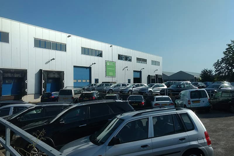 Voller Parkplatz vor weißer Lagerhalle mit blauen Toren und zahlreichen geparkten Autos, Zaun im Vordergrund.