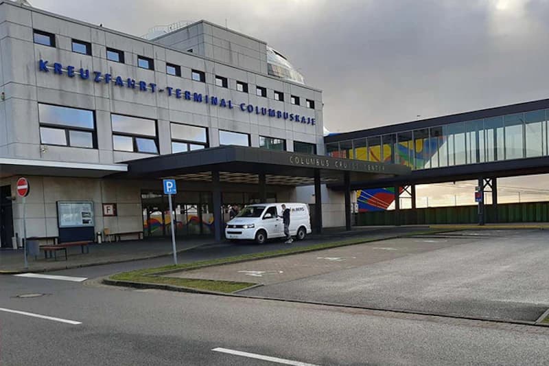 KREUZFAHRT-TERMINAL COLUMBUSKAJE mit Vordach, weißem Transporter auf der Vorfahrt und regennassem Parkplatz.