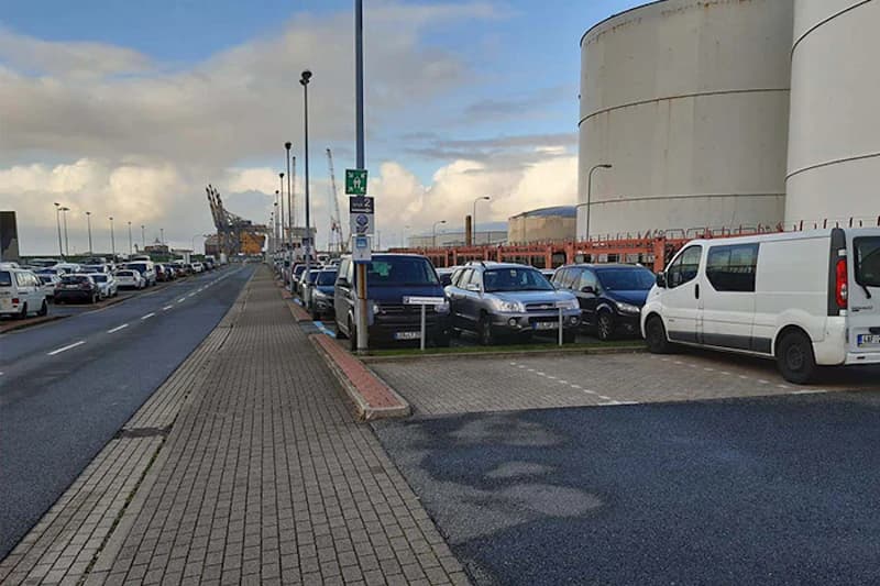 Gehweg und Straße neben Freigelände mit geparkten Autos, großen Lagerbehältern und Kränen am Hafen.