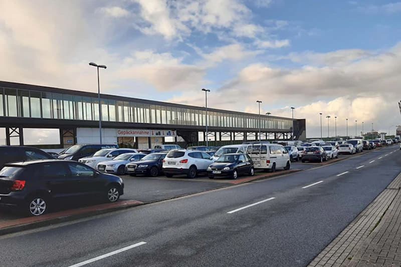 Parkplatz mit zahlreichen geparkten Autos neben erhöhter verglaster Gangway und Gebäude mit Schild Gepäckannahme.