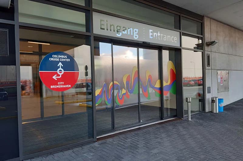 Schiebetür-Eingang mit buntem Wellenaufkleber und runder Columbus Cruise Center Tafel am Hafengebäude in Bremerhaven.