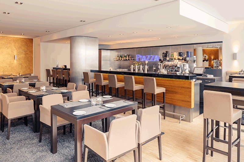Helle Hotelbar mit Theke, hohen Barhockern, mehreren Esstischen und beigen Polsterstühlen sowie Neonbeschriftung PIAZZA.