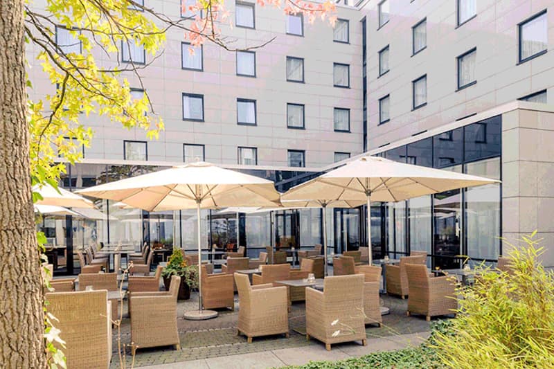 Hotelterrasse mit Flechtstühlen, Tischen und Sonnenschirmen vor mehrstöckigem Hotelgebäude.