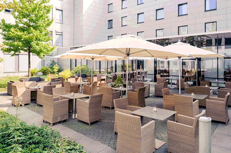 Hotelterrasse mit Tischen, Rattanstühlen, Sonnenschirmen und Gebäudefassade im Hintergrund.