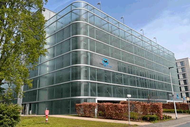 Mehrstöckiges Parkhaus mit glasverkleideter Fassade und FRA Parking Logo, Hecken und Gehweg im Vordergrund.