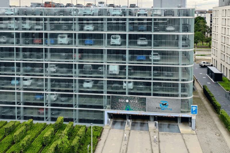 Mehrstöckiges Parkhaus mit gläserner Fassade, sichtbar geparkte Autos und drei Einfahrtrampen, Hecken im Vordergrund.