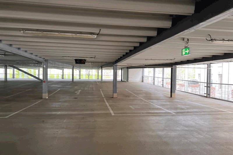 Leeres Parkdeck im mehrstöckigen Parkhaus mit markierten Stellplätzen, Stahlstützen und grünem Notausgangsschild.