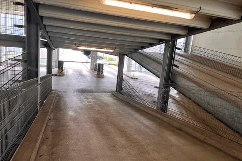 Abwärtsrampe in einem mehrstöckigen Parkhaus mit Metallträgern, Drahtgitterseiten und Schranke am Ausgang.