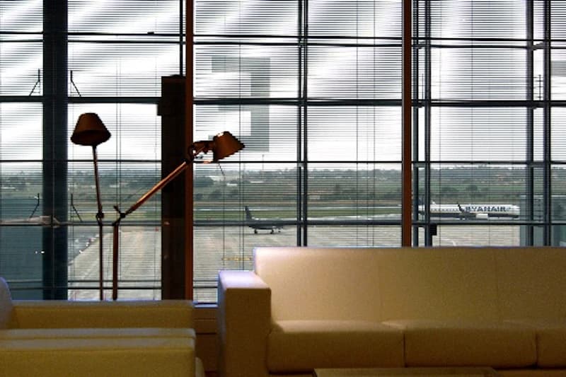 Wartebereich mit Sofas und Leselampen vor großen Glasfenstern, Blick auf Rollfeld mit Flugzeugen.