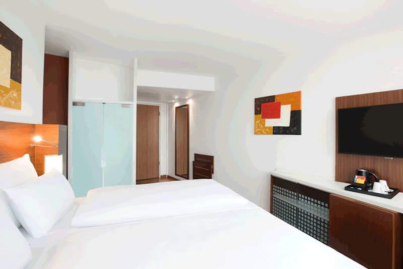 Hotelzimmer mit zwei Betten, weißer Bettwäsche, Holz-Kopfteil, Nachttischlampe und Glastür zum Bad.