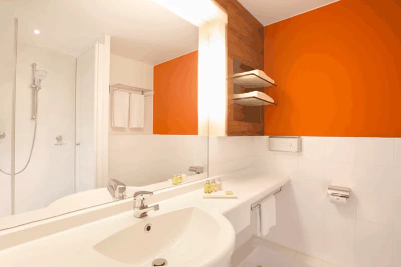 Badezimmer mit Waschbecken, großem Spiegel, oranger Wand, Duschkabine, Handtüchern und kleinen Toilettenartikeln.