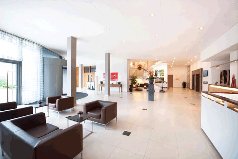 Helle Hotellobby mit braunen Sesseln, Rezeption rechts und Treppe im Hintergrund.
