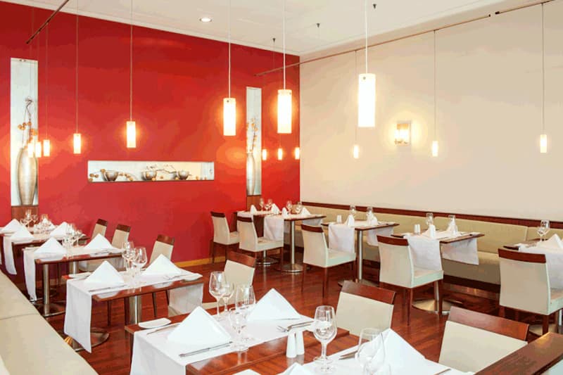 Restaurant mit weißen Tischdecken, gefalteten Servietten, Weingläsern, roter Akzentwand und hängenden Lampen.