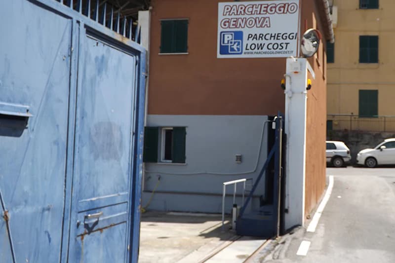 Geöffnete blaue Metalltore links, Einfahrt mit Schranke und Gebäude mit Schild Parcheggio Genova am Hafen.