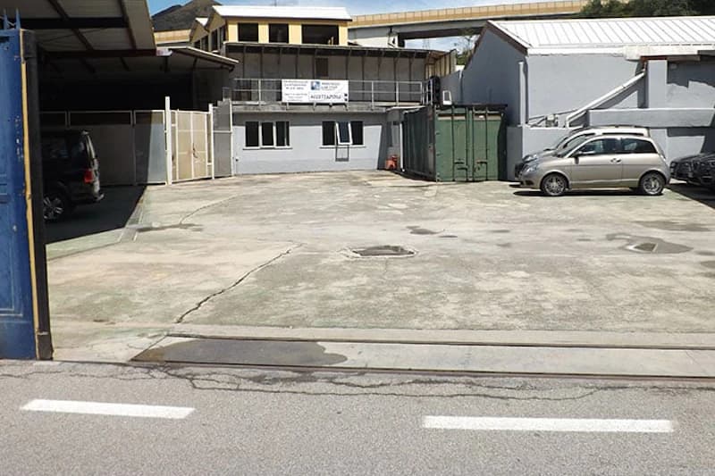 Asphaltierter Parkplatz mit kleinem grauem Gebäude, zwei Autos rechts, Seecontainer und geöffnete Torzufahrt.