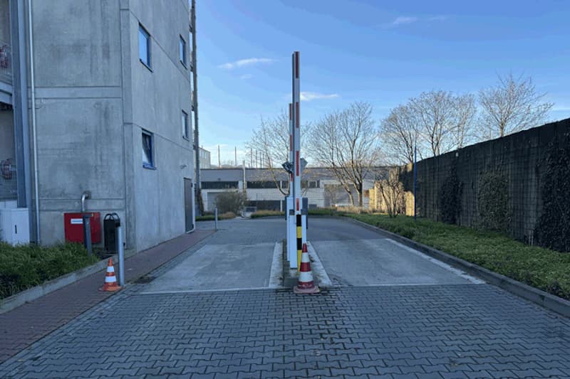 Zufahrt mit Schrankenanlage zu Parkhaus zwischen Betongebäude links und begrüntem Zaun rechts.