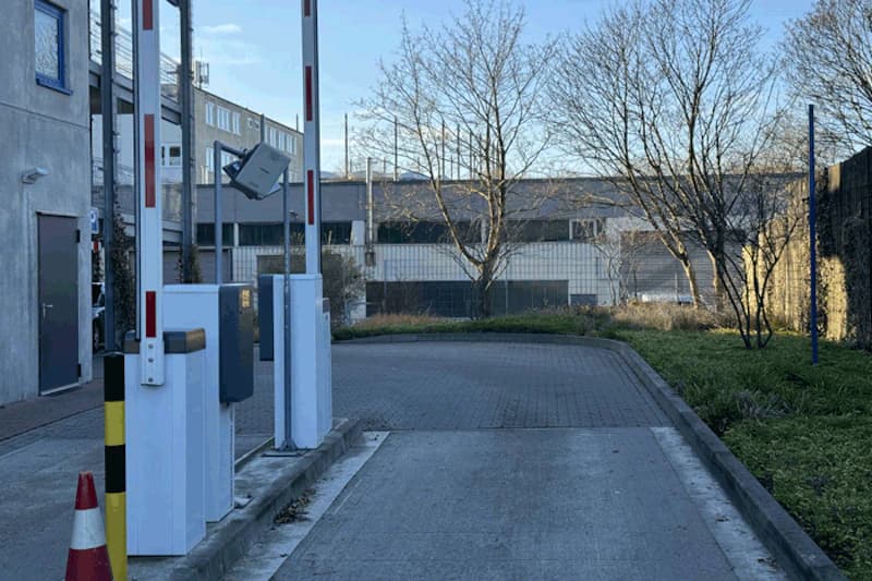 Zufahrt mit Schranken, Ticketgeräten und Überwachungskamera vor umzäuntem Hof und Gebäuden.