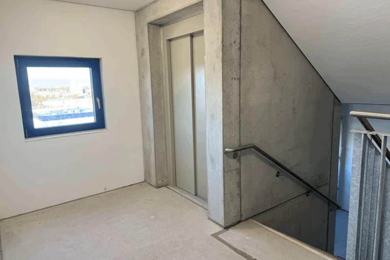 Treppenhausgeschoss mit geschlossener Aufzugtür, Betonwänden, Handlauf und blau gerahmtem Fenster.