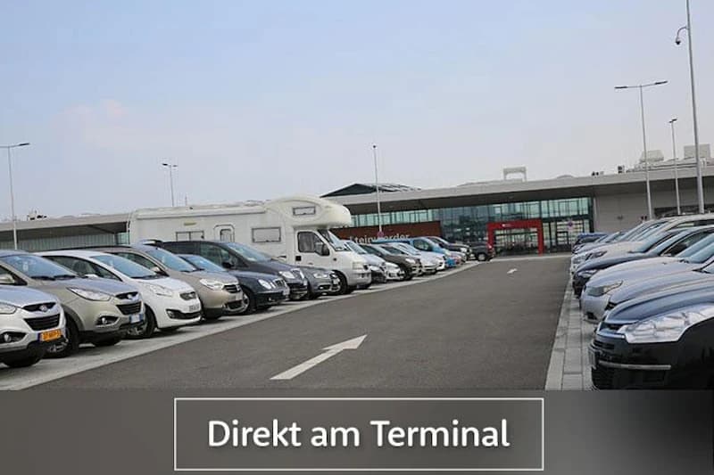 Parkplatz mit zwei Reihen geparkter Autos und einem Wohnmobil, Asphaltweg Richtung Terminalgebäude.