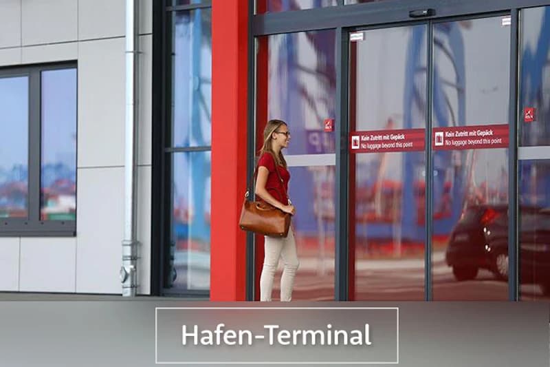 Frau mit brauner Tasche steht vor gläserner Schiebetür mit rotem Rahmen und Hafenkränen in der Spiegelung.