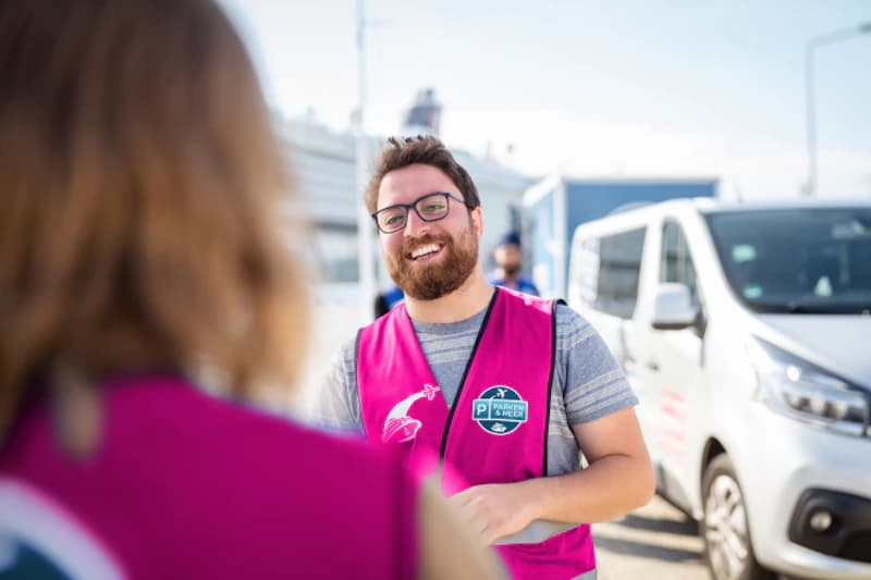 Mann mit rosa Sicherheitsweste und Parken & Meer Logo lächelt vor einem weißen Transporter am Hafen.