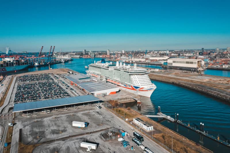 Kreuzfahrtschiff AIDAluna am Rostocker Hafen neben großem Parkplatz und Terminal, Hafenkräne im Hintergrund.