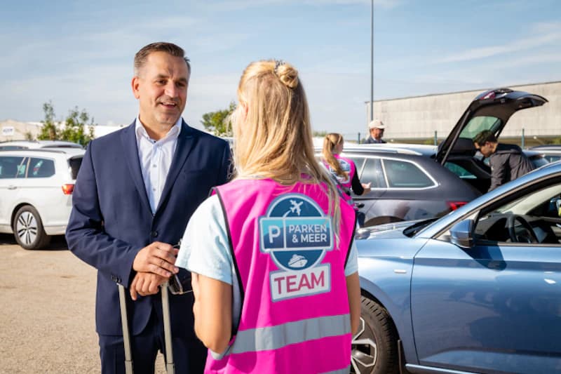 Mann im Anzug mit Trolley spricht auf Parkplatz mit Mitarbeiterin in pinker Parken & Meer Team-Weste, Autos im Hintergrund.