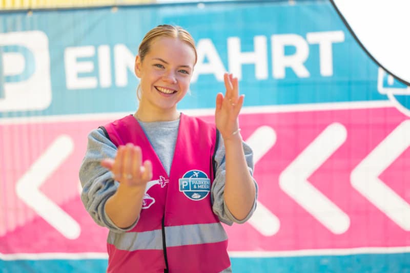 Lächelnde Parkplatzmitarbeiterin in pinker Weste mit Parken & Meer-Logo vor Schild Einfahrt, winkend.