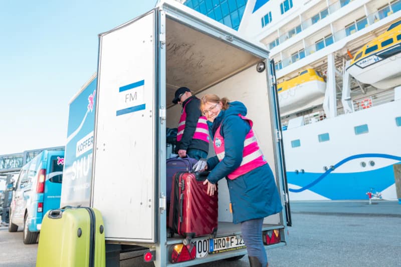 Zwei Mitarbeiter in rosa gestreiften Westen laden Koffer aus offenem Anhänger vor Kreuzfahrtschiff.