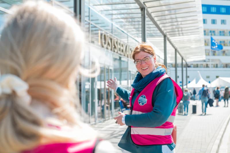 Frau in pinker Warnweste zeigt lächelnd auf den Eingang des Ostseekai-Terminals, im Vordergrund unscharfe Person.