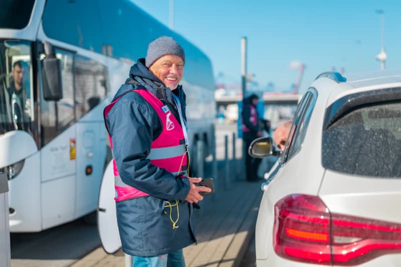 Mitarbeiter in pinker Warnweste lächelnd neben weißem Auto und Reisebus am Hafen.