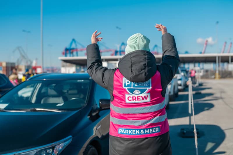 Person in rosa Warnweste mit Aufdruck leitet Autos auf Parkfläche am Hafen Rostock an.