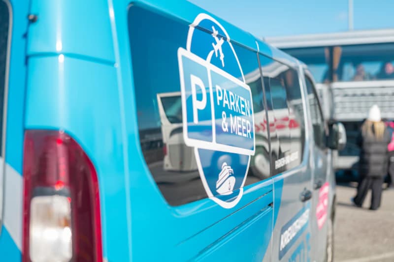 Blauer Transferbus mit Parken & Meer-Logo, reflektierende Seitenfläche, Bussen und wartenden Personen im Hafen.