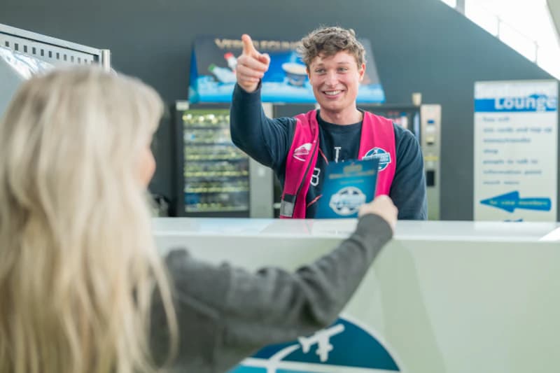 Mitarbeiter am Empfangstresen reicht einer Kundin eine Karte, im Hintergrund Getränkeautomat und Hinweisschild.