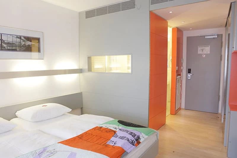 Kleines Hotelzimmer mit zwei Einzelbetten, buntem Überwurf, hellen Wänden, Holzfußboden und offener Zimmertür.