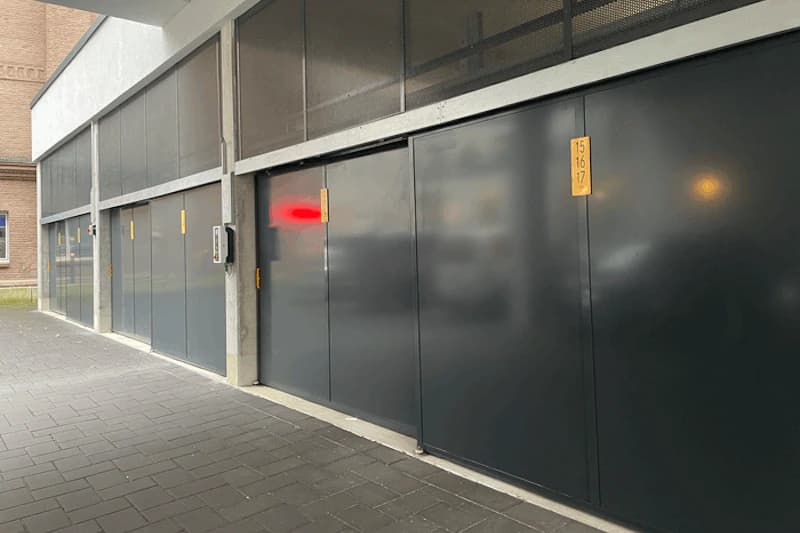 Reihe geschlossener dunkelgrauer Garagentore mit Nummerntafeln und Gegensprechanlage unter einem Gebäude.