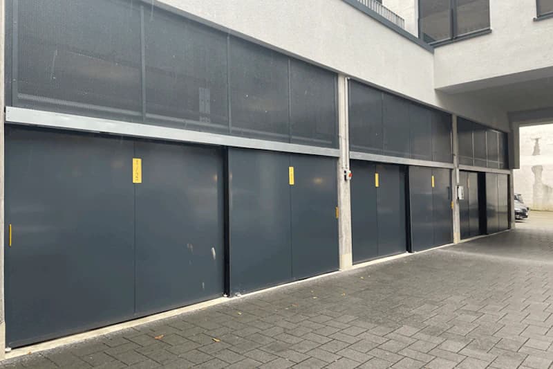 Reihe geschlossener dunkelgrauer Garagentore unter einem Gebäude mit gepflasterter Zufahrt.