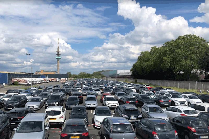 Umzäuntes Parkplatzgelände am Hafen Köln mit dichtstehenden Pkw und Sendeturm am Horizont.