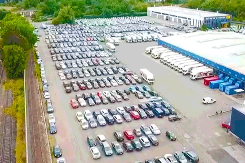 Großer Parkplatz mit vielen geparkten Autos in Reihen neben Lagerhalle und Gleisen.