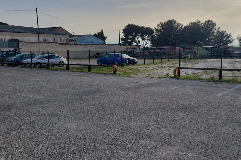 Gepflasterter umzäunter Parkplatz mit wenigen geparkten Autos und Metallzaun, Gebäude und Bäume im Hintergrund.