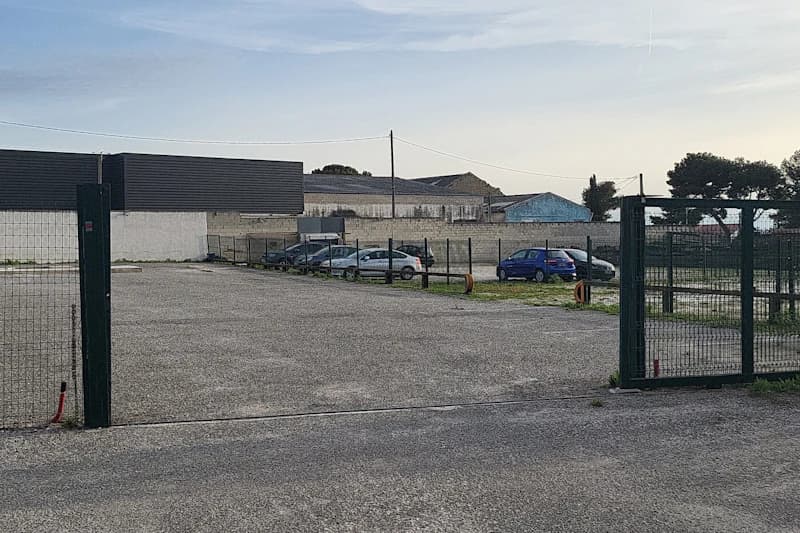 Eingezäunter Parkplatz mit offenem Tor, Asphaltfläche und wenigen geparkten Autos vor Lagergebäuden.