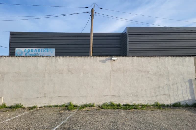 Leerer Parkstreifen mit weißen Markierungen vor einer hohen Betonmauer, dahinter graue Gebäude, Strommast und Überwachungskamera.