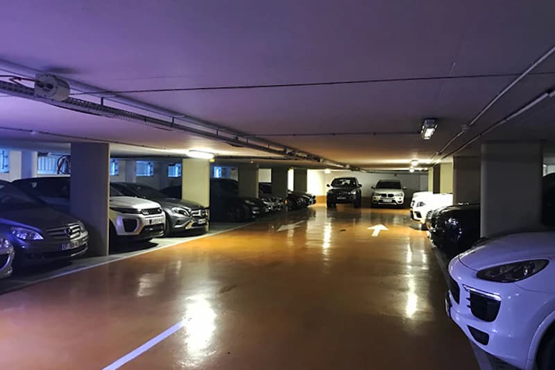 Tiefgarage mit geparkten Autos in Reihen, Stützpfeilern, Deckenbeleuchtung und Richtungspfeil auf dem Boden.