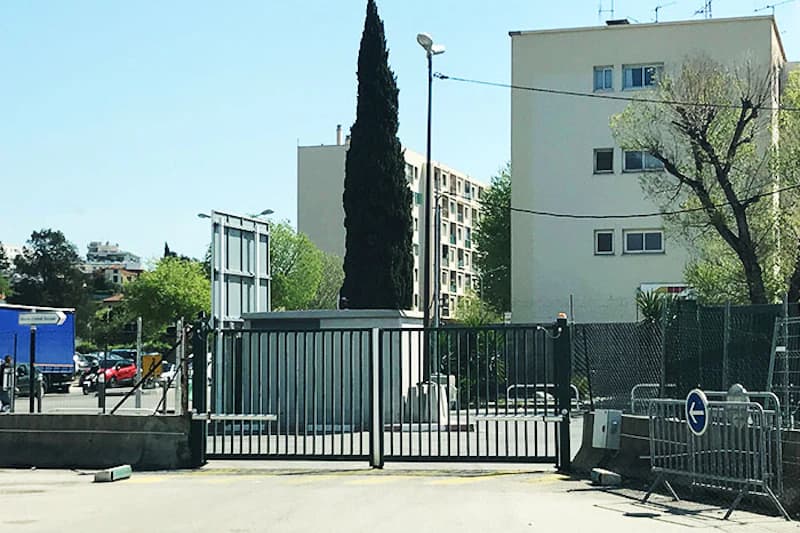 Geschlossene Metallzufahrt mit Tor, umzäuntem Bereich, Wachhäuschen und Wohngebäuden im Hintergrund.